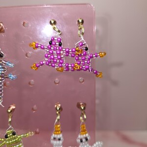 May include: A pair of beaded earrings featuring two pink, grid-patterned frog figures with black eyes and orange feet, suspended from gold-tone metal studs. The figures are connected by a thin white thread, creating a cohesive, playful design.