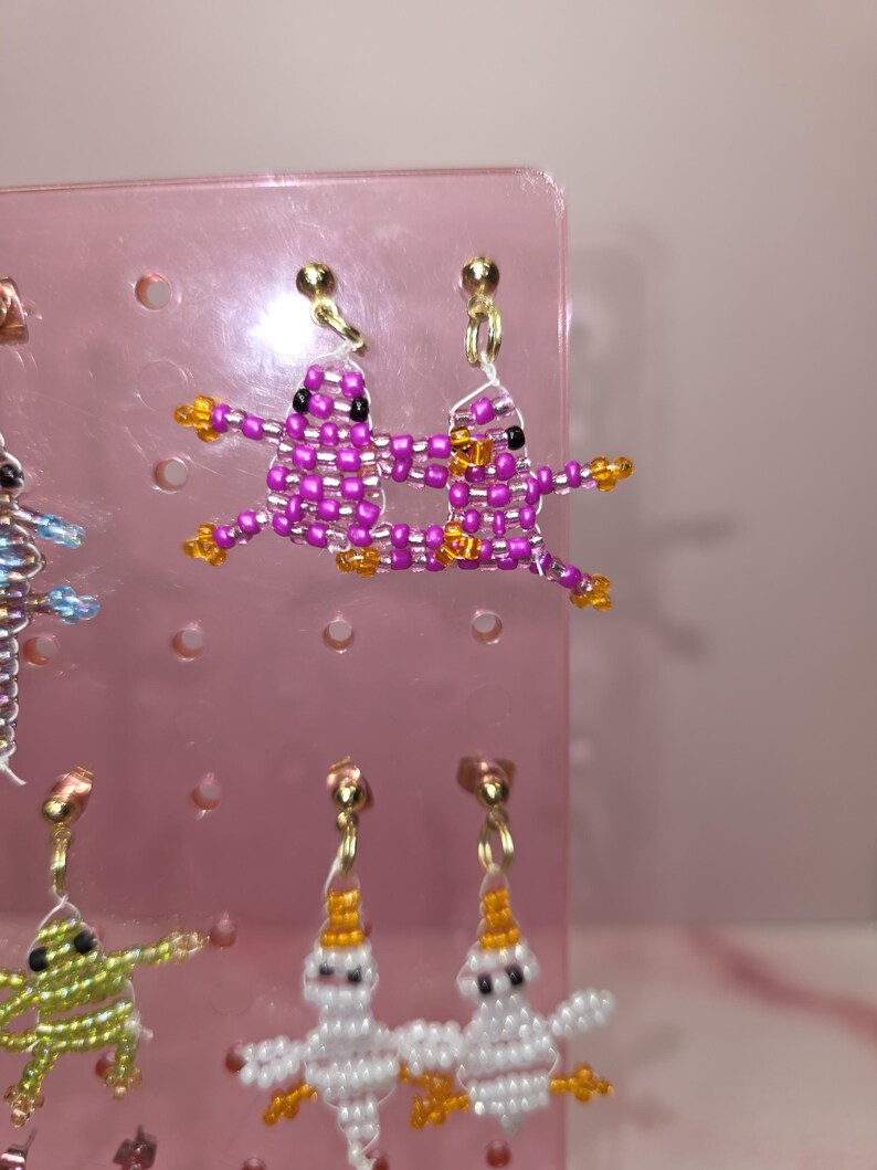 May include: A pair of beaded earrings featuring two pink, grid-patterned frog figures with black eyes and orange feet, suspended from gold-tone metal studs. The figures are connected by a thin white thread, creating a cohesive, playful design.