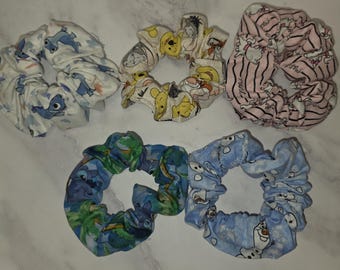 May include: Five fabric hair scrunchies featuring various cartoon character prints. The collection includes a white scrunchie with blue creatures, a beige one with yellow bears, a pink striped one with white cats, a blue one with green foliage, and a light blue one with snowmen.