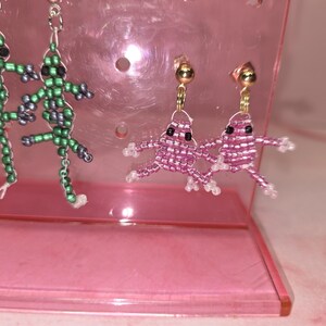 May include: Two pairs of beaded earrings. One pair features green lizard-shaped figures with black eyes and silver-tone hooks. The second pair features pink frog-shaped figures with black eyes and gold-tone stud posts. The earrings are displayed on a pink stand.