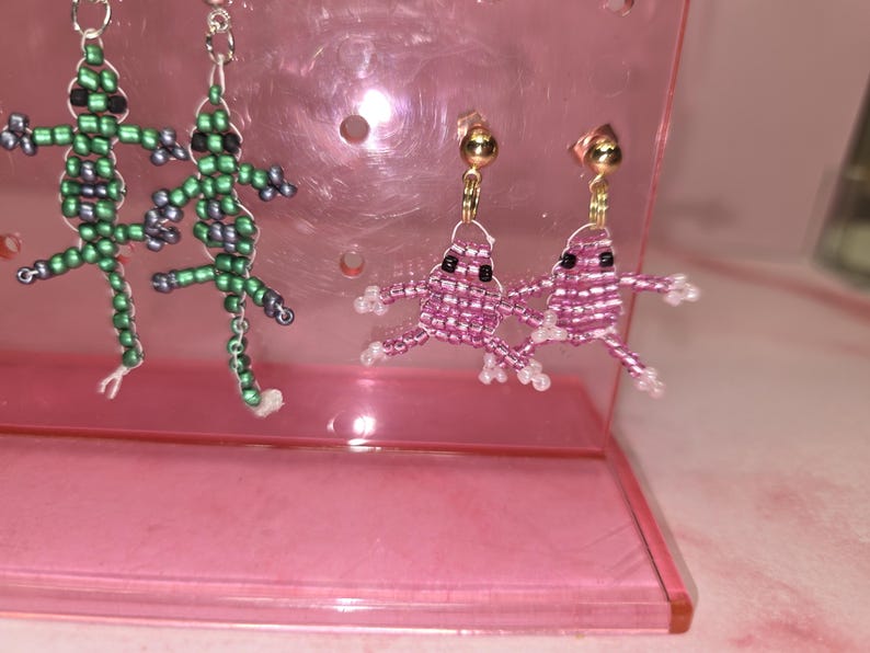 May include: Two pairs of beaded earrings. One pair features green lizard-shaped figures with black eyes and silver-tone hooks. The second pair features pink frog-shaped figures with black eyes and gold-tone stud posts. The earrings are displayed on a pink stand.