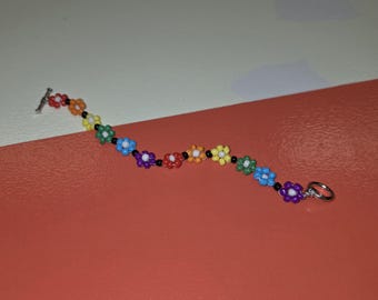 May include: A beaded bracelet featuring a series of small, multi-colored flower-shaped beads in red, orange, yellow, green, blue, and purple, separated by tiny black spacer beads, finished with a silver-toned metal toggle clasp closure.