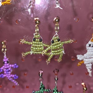 May include: A pair of stud earrings featuring small, handmade beaded frogs in shades of lime green with black bead eyes. The frogs are constructed from tiny glass seed beads and are attached to gold-tone metal posts. Other beaded animal charms are visible.