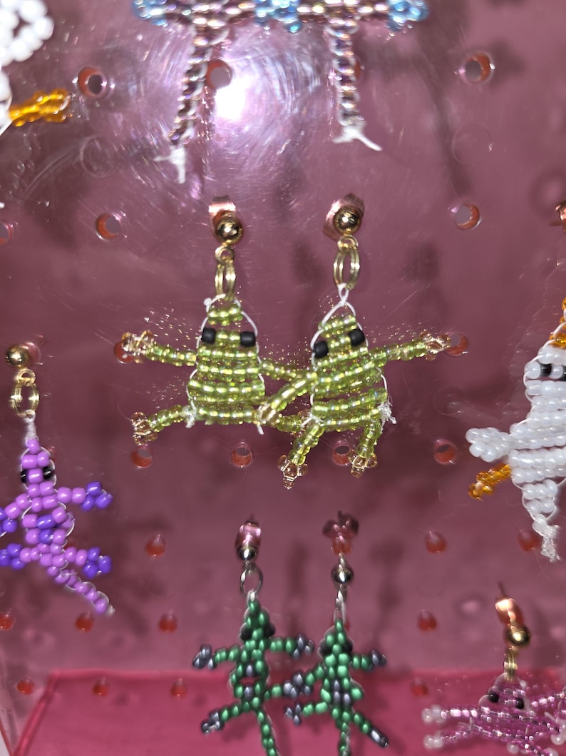 May include: A pair of stud earrings featuring small, handmade beaded frogs in shades of lime green with black bead eyes. The frogs are constructed from tiny glass seed beads and are attached to gold-tone metal posts. Other beaded animal charms are visible.