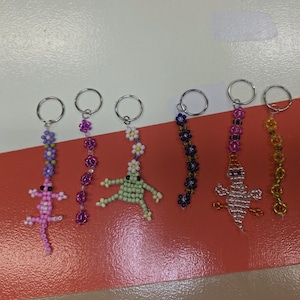 May include: Six beaded keychains featuring various designs including a pink lizard, a green frog, a white ghost, and three floral strands. Each keychain is constructed from small colourful glass beads and attached to a silver-toned metal ring.