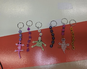May include: Six beaded keychains featuring various designs including a pink lizard, a green frog, a white ghost, and three floral strands. Each keychain is constructed from small colorful glass beads and attached to a silver-toned metal ring.