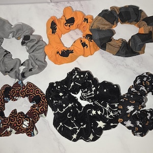 May include: A collection of six fabric hair scrunchies featuring various patterns including a grey bat print, an orange cat print, a black and orange plaid, a brown geometric pattern, a black star constellation print, and a black ghost print.