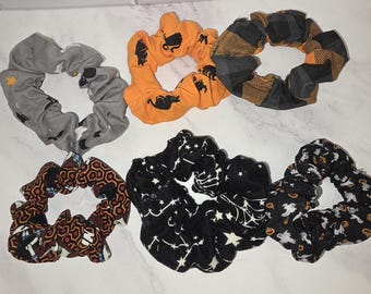 May include: A collection of six fabric hair scrunchies featuring various patterns including a grey bat print, an orange cat print, a black and orange plaid, a brown geometric pattern, a black star constellation print, and a black ghost print.