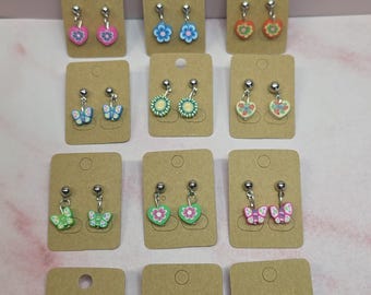 May include: Twelve pairs of dangle earrings displayed on individual brown cardboard cards. The earrings feature colorful charms in shapes of hearts, flowers, butterflies, and citrus slices, each attached to a silver-toned metal post backing.