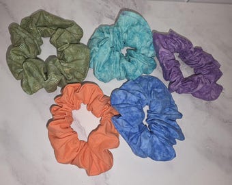 May include: A set of five fabric hair scrunchies in various colors and textures. The collection includes a green one with a wavy line pattern, a solid orange one, a mottled turquoise one, a mottled blue one, and a mottled purple hair accessory.