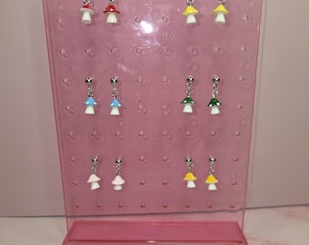 May include: A collection of six pairs of mushroom-shaped dangle earrings displayed on a translucent pink plastic stand. The earrings feature small, colorful mushroom caps in red, yellow, blue, green, pink, and orange, each with a white stem.