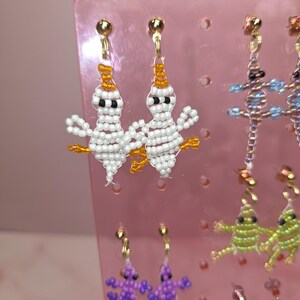 May include: A pair of white beaded ghost earrings with orange accents and black eyes, hanging from gold-tone posts. These earrings are displayed on a pink plastic stand alongside other beaded lizard-shaped earrings in purple, green, and blue.