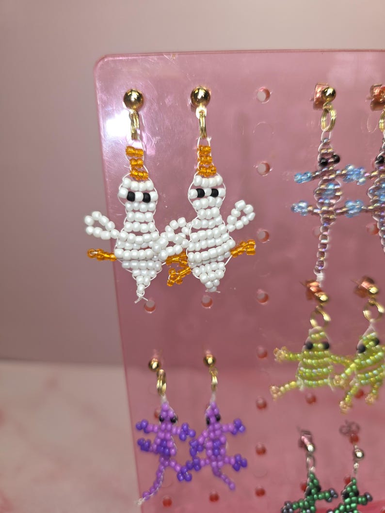 May include: A pair of white beaded ghost earrings with orange accents and black eyes, hanging from gold-tone posts. These earrings are displayed on a pink plastic stand alongside other beaded lizard-shaped earrings in purple, green, and blue.