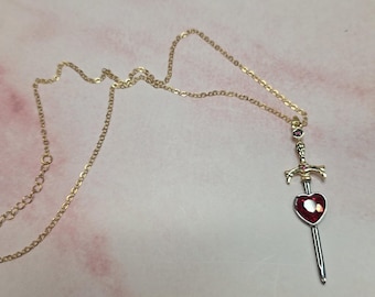 May include: A gold-toned chain necklace featuring a pendant shaped like a sword. The sword hilt is gold-toned with small pink accents, and the blade is silver-toned, featuring a prominent, faceted, heart-shaped red gemstone at the center.