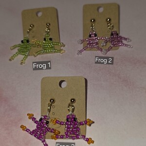 May include: Three pairs of beaded frog earrings displayed on brown cardboard cards. Frog 1 features green beads, Frog 2 features pink beads, and Frog 3 features purple beads with orange accents. Each pair includes gold-toned stud earring hardware.