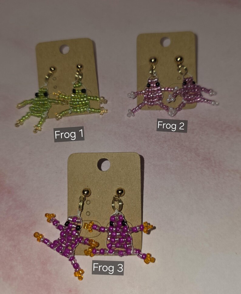 May include: Three pairs of beaded frog earrings displayed on brown cardboard cards. Frog 1 features green beads, Frog 2 features pink beads, and Frog 3 features purple beads with orange accents. Each pair includes gold-toned stud earring hardware.
