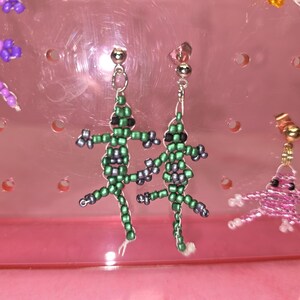 May include: A pair of dangle earrings featuring lizard-shaped charms crafted from small green and grey seed beads. Each lizard has black bead eyes and is attached to a silver-toned metal post backing. The earrings are displayed on a pink stand.