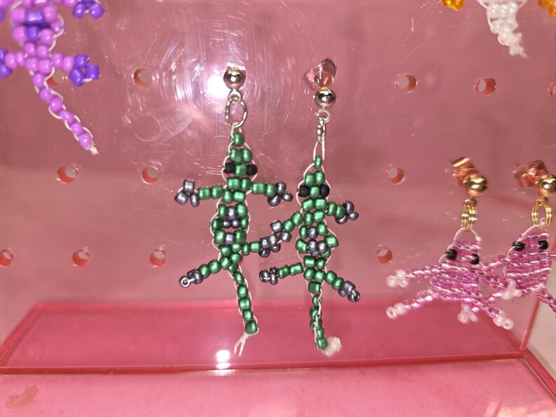 May include: A pair of dangle earrings featuring lizard-shaped charms crafted from small green and grey seed beads. Each lizard has black bead eyes and is attached to a silver-toned metal post backing. The earrings are displayed on a pink stand.