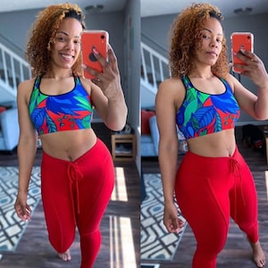 May include: A woman wearing a red sports bra with a blue, green and red floral print and matching red leggings. She is holding a phone in her right hand and smiling at the camera.