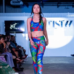 May include: A woman is modelling a blue, red and green patterned sports bra and leggings. The bra and leggings have a tropical leaf design. The woman is wearing black high heels and has her hair pulled back in a braid.