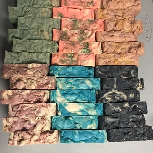 May include: A collection of handmade soap bars in various colours and patterns. The bars are stacked in three rows, with each row featuring a different colour scheme. The top row features green and white bars, the middle row features pink and white bars, and the bottom row features blue and white bars, black and white bars.