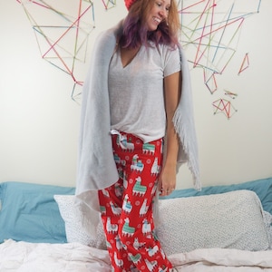 Può includere: Una donna indossa pantaloni da pigiama rossi con stampa di lama, una maglietta bianca, una coperta grigia e un berretto di lana rosso. I pantaloni hanno un tema natalizio con fiocchi di neve e altri motivi festivi.