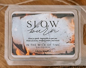 Puede incluir: Un recipiente de plástico transparente que contiene una cera perfumada de soja. La etiqueta dice "SLOW burn" con el texto "IN THE WICK OF TIME CALIFORNIA" y "3 Oz Soy Wax Melt". Las notas de fragancia incluyen cacao, ámbar ahumado y almizcle.