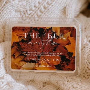 May include: A clear plastic container of soy wax melt, with the text "THE 'BER months" and "IN THE WICK OF TIME CALIFORNIA". The container is placed on a cream-colored cable knit sweater. The wax melt has a fall leaves design.