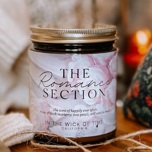 May include: A glass jar candle with a gold lid and a label that reads "The Romance Section." The label features a floral design with the text "The scent of happily ever afters." The candle is on a wooden surface.