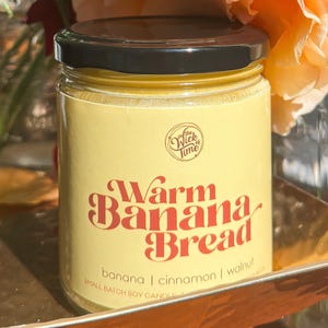 May include: A glass jar candle with a black lid and a yellow label. The label reads "Warm Banana Bread" in a retro font, with the scent ingredients listed below. The candle is on a gold tray, with a peach rose in the background.