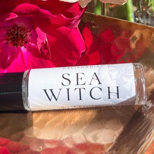 Puede incluir: Un frasco de bola de cristal transparente con tapa negra, etiquetado "SEA WITCH" en texto negro. El frasco descansa sobre una superficie dorada, con vibrantes flores rojas y melocotón en el fondo.