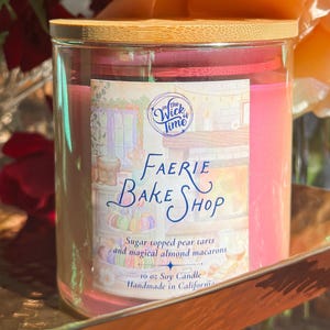 Puede incluir: Un tarro de vela de vidrio con tapa de madera. La etiqueta dice "Faerie Bake Shop" con texto sobre tartas de pera y macarons de almendras. La vela es rosa y pesa 283 gramos. Hecho a mano en California.