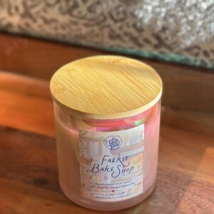 May include: A glass candle jar with a wooden lid. The candle is pink and has a label that reads "Faerie Bake Shop". The label also mentions "sugar topped pear tarts and magical almond macarons".
