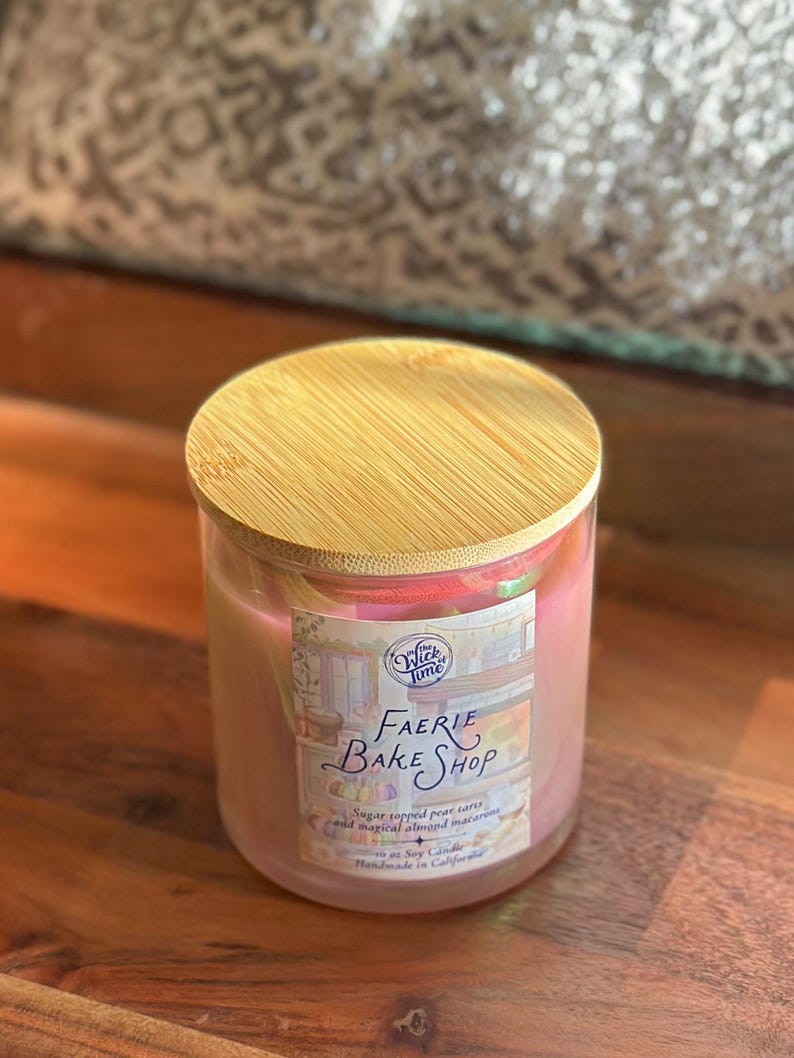 May include: A glass candle jar with a wooden lid. The candle is pink and has a label that reads "Faerie Bake Shop". The label also mentions "sugar topped pear tarts and magical almond macarons".