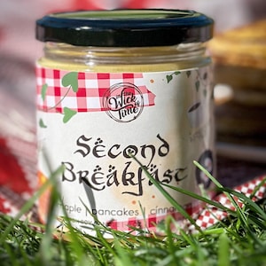 Puede incluir: Un tarro de vidrio con tapa negra lleno de una crema amarilla. La etiqueta es blanca con detalles rojos y verdes y dice "In the Wick of Time Second Breakfast Maple Pancakes Cinnamon".