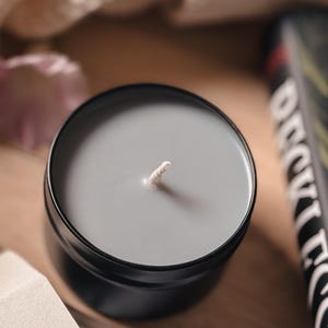 Puede incluir: Una vela gris en un recipiente de hojalata negro con una mecha blanca. La vela est&aacute; sobre una superficie de madera.