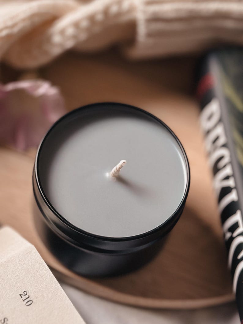 Puede incluir: Una vela gris en un recipiente de hojalata negro con una mecha blanca. La vela est&aacute; sobre una superficie de madera.