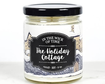 May include: A glass jar candle with a black lid. The label features a winter cottage scene with the text "IN THE WICK OF TIME" and "The Holiday Cottage". The candle is vegan and soy, with a net weight of 8 oz.
