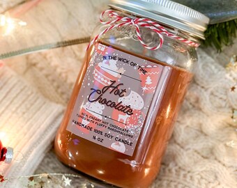 May include: A glass jar candle filled with brown wax, labeled "Hot Chocolate." The label includes the text "In the Wick of Time," and "Rich, creamy hot chocolate topped with fluffy marshmallows." The candle is 16 oz and tied with red and white twine.