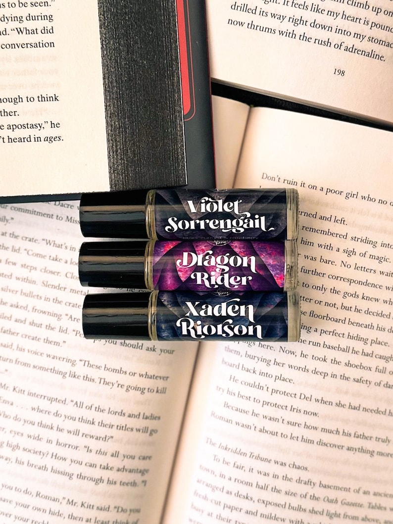 Puede incluir: Tres botellas de vidrio con rodillo con etiquetas que presentan nombres de personajes de fantas&iacute;a. Las etiquetas est&aacute;n en un esquema de color morado, rojo y negro. Los nombres en las etiquetas son Violet Sorrengail, Dragon Rider y Xaden Riotson.