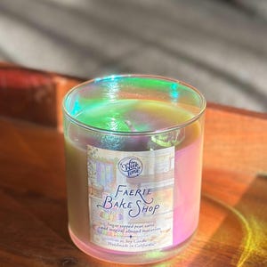 May include: A glass candle jar with a pearlescent sheen. The candle has a label that reads "Faerie Bake Shop" and "Sugar topped pear tarts and magical almond macarons." The candle is 10 oz and handmade in California.