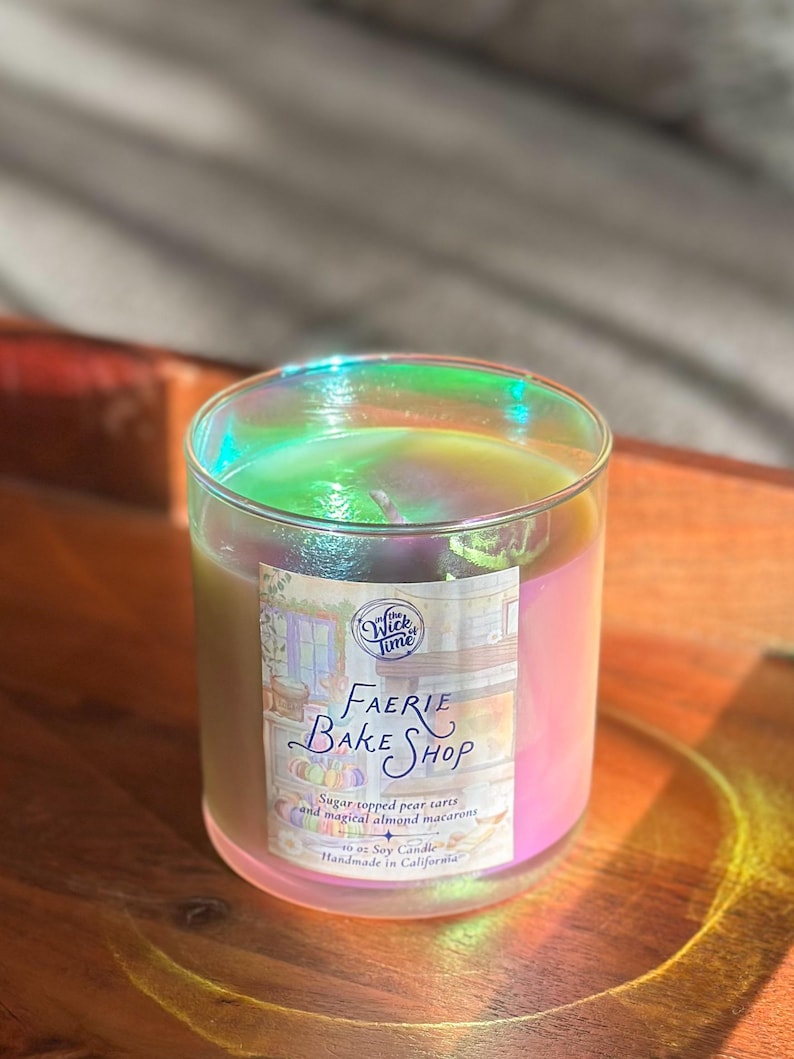 May include: A glass candle jar with a pearlescent sheen. The candle has a label that reads "Faerie Bake Shop" and "Sugar topped pear tarts and magical almond macarons." The candle is 10 oz and handmade in California.