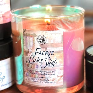 May include: A lit soy candle in a clear glass jar. The label reads "Faerie Bake Shop" with text about pear tarts and almond macarons. The candle is 10 oz.