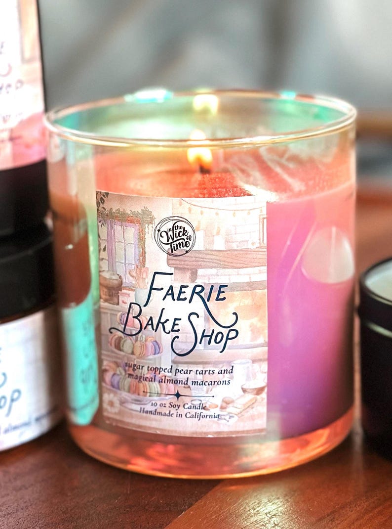 May include: A lit soy candle in a clear glass jar. The label reads "Faerie Bake Shop" with text about pear tarts and almond macarons. The candle is 10 oz.