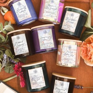 May include: An assortment of seven scented soy candles in glass jars with wooden lids. Each candle has a label with a unique name, such as "Reading Nook" and "Twilight Tavern." The candles are arranged on a brown surface with flowers and a book.