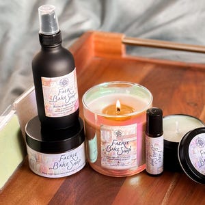 May include: A collection of Faerie Bake Shop products, including a black spray bottle, a jar, a candle in a glass, a roller bottle, a small tin, and wax melts. The products are arranged on a wooden tray. The labels have the text "Faerie Bake Shop".