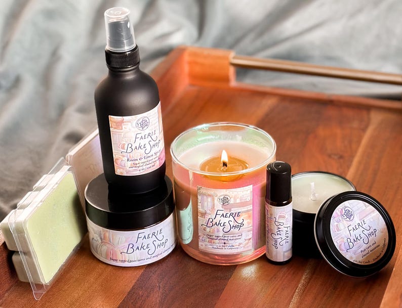 May include: A collection of Faerie Bake Shop products, including a black spray bottle, a jar, a candle in a glass, a roller bottle, a small tin, and wax melts. The products are arranged on a wooden tray. The labels have the text "Faerie Bake Shop".