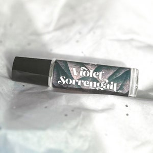 Puede incluir: Un frasco de vidrio transparente con rodillo y tapa negra. La etiqueta es un dise&ntilde;o abstracto verde y morado con el texto "Violet Sorrengail" en blanco.
