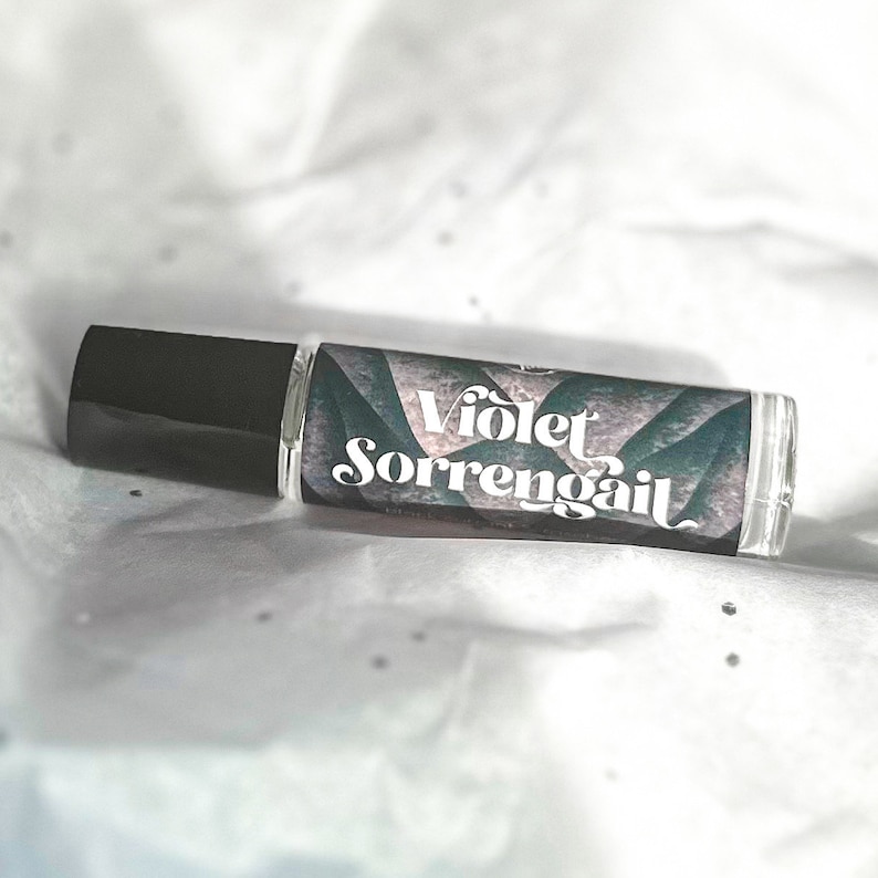 Puede incluir: Un frasco de vidrio transparente con rodillo y tapa negra. La etiqueta es un dise&ntilde;o abstracto verde y morado con el texto "Violet Sorrengail" en blanco.