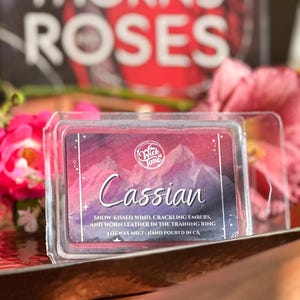 May include: A clear plastic container holding a wax melt. The label features the name "Cassian" over a mountain scene. The text reads "Snow-kissed wind, crackling embers, and worn leather in the training ring." The product is 3 oz.
